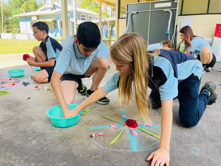 Two male students are reaching into a green bowl of sand to add more colour to their symetical, circular Rangoli pattern half created on the concrete in front of them. Other students can be seen in the background, also creating Rangoli.