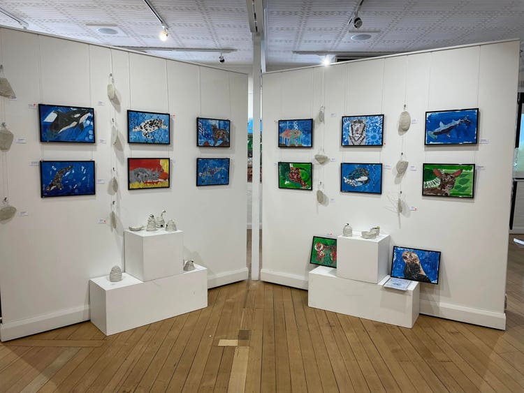 Two white Gallery divider walls meet at an angle. On the wall are 14 colourful mosaics of endangered animals in black frames. In front of the wall display are white stage display boxes with white airdried clay figures of beehives and bees.